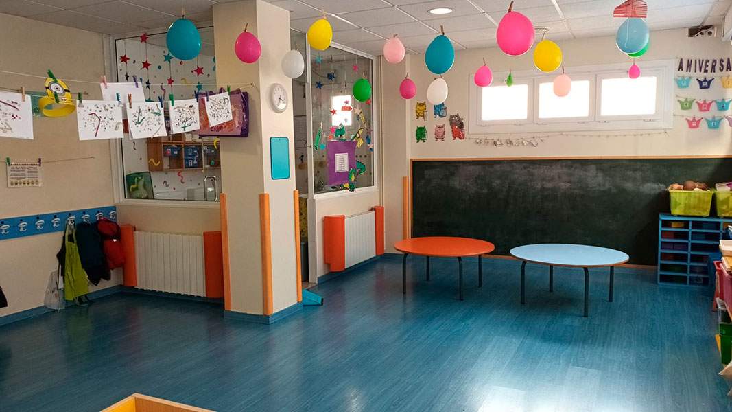Imagen de aula para niños de 24 a 36 meses, luminosa, con mesas y juegutes, decorada con dibujos y globos.