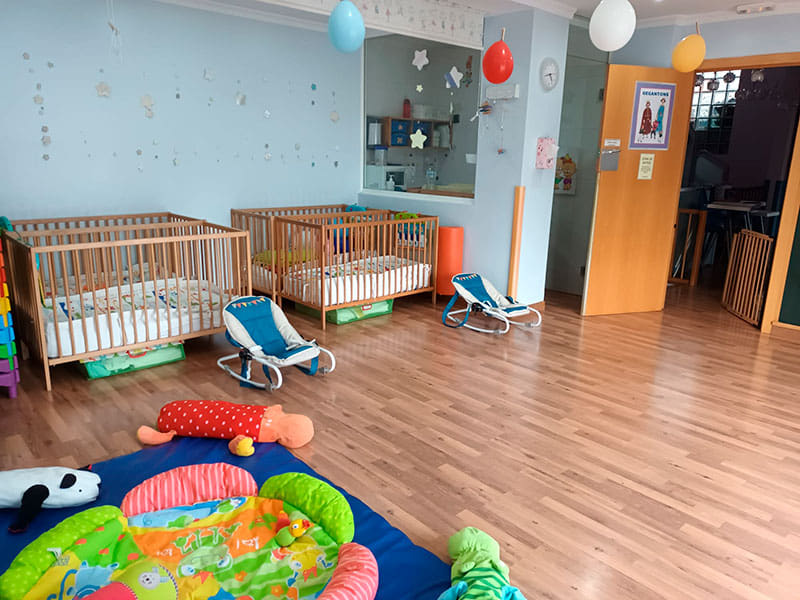 Imagen de aula para niños de 0 a 12 meses con mantas juguetes en el suelo y cunas pegadas a la pared.